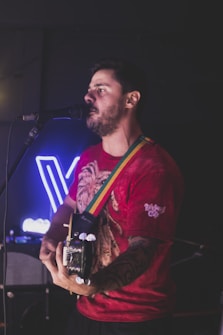 A man is playing an electric guitar while singing into a microphone. He has short dark hair and a beard and is wearing a red t-shirt with a graphic design and a colorful strap on the guitar. The background is dimly lit with a blue neon sign.