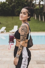 A young woman with a ponytail is standing near a pool, holding a beverage container. She is wearing a black sports bra and black and white track pants, displaying detailed tattoos on her arm and shoulder. The setting is outdoors, with a grassy area and lounge chairs visible in the background.