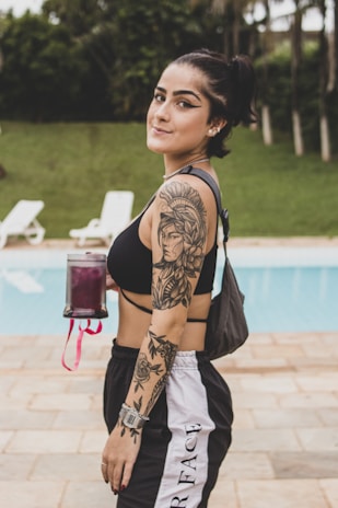 A young woman with a ponytail is standing near a pool, holding a beverage container. She is wearing a black sports bra and black and white track pants, displaying detailed tattoos on her arm and shoulder. The setting is outdoors, with a grassy area and lounge chairs visible in the background.