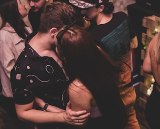 Several people are socializing closely in dim lighting, likely in a party or club setting. Two individuals in the foreground are embracing each other, creating a sense of intimacy. Other people are visible in the background, engaging in conversation and activities. The environment appears lively with casual outfits being worn by the individuals.