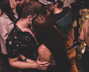 Several people are socializing closely in dim lighting, likely in a party or club setting. Two individuals in the foreground are embracing each other, creating a sense of intimacy. Other people are visible in the background, engaging in conversation and activities. The environment appears lively with casual outfits being worn by the individuals.