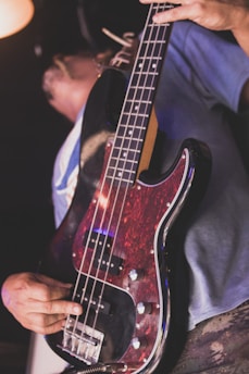 A person holding and playing an electric bass guitar with a dark red and black body. The bass guitar features silver knobs and pickups. The focus is on the guitar and the person’s hand, with the background out of focus.