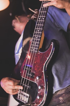 A person holding and playing an electric bass guitar with a dark red and black body. The bass guitar features silver knobs and pickups. The focus is on the guitar and the person’s hand, with the background out of focus.
