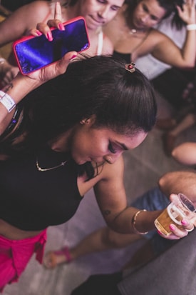 A young woman with long dark hair is dancing while holding a drink in one hand and a phone in the other. She appears to be in a festive or party setting with other people in the background. She is wearing a sleeveless black top and pink shorts, and there are bracelets on her wrist.