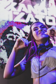A woman is smiling and dancing, wearing a sparkling top and headphones around her neck. She has vibrant makeup and purple highlights in her hair. The background features a colorful, brightly lit digital screen.