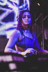 A woman wearing headphones is focused on her task at a DJ booth, with a colorful LED screen in the background casting vibrant blue and purple lights. Her tattoos and casual attire contribute to a modern, artistic vibe.