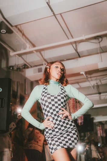 A person poses confidently with hands on hips, wearing a checkered dress layered over a light blue long-sleeve top. The setting appears to be indoors with exposed piping on the ceiling and soft lighting, suggesting a fashion or social event.
