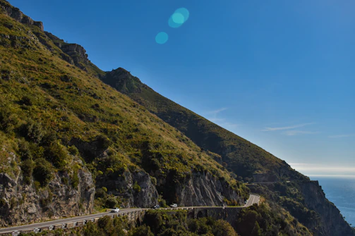 A winding coastal road hugging cliffs with turquoise waves crashing below under a clear blue sky.
