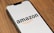 A smartphone displaying the Amazon logo against a plain background, positioned at an angle on a wooden surface.