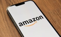 A smartphone displaying the Amazon logo against a plain background, positioned at an angle on a wooden surface.