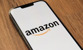 A smartphone displaying the Amazon logo against a plain background, positioned at an angle on a wooden surface.