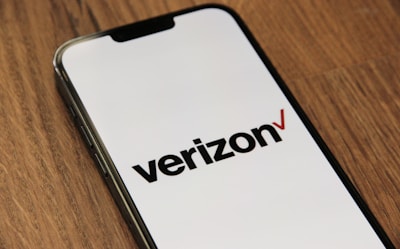 A smartphone lies on a wooden surface displaying the Verizon logo on its screen. The phone is positioned at an angle, showcasing the logo prominently against a white background.
