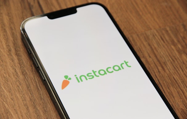 A smartphone is placed on a wooden surface displaying the Instacart logo on its screen. The logo consists of the word 'instacart' in green, accompanied by a carrot icon to the left.