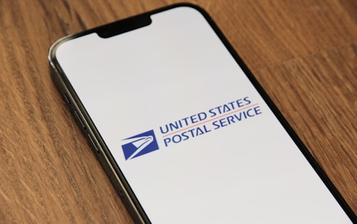 A smartphone lies on a wooden surface with its screen displaying the United States Postal Service logo. The logo is prominently featured on a white background.