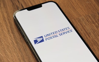 A smartphone lies on a wooden surface with its screen displaying the United States Postal Service logo. The logo is prominently featured on a white background.