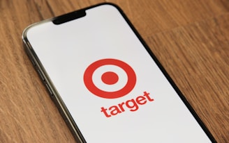 A smartphone displays the Target logo, featuring a red bullseye and the word 'target' in red, on its screen. The phone is placed on a wooden surface.