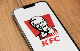 A smartphone is placed on a wooden surface, displaying the KFC logo. The logo consists of the image of Colonel Sanders in black and white against a red background, with the letters 'KFC' beneath it.