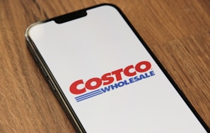 A smartphone displaying the Costco Wholesale logo on its screen, placed on a wooden surface.