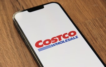 A smartphone displaying the Costco Wholesale logo on its screen, placed on a wooden surface.