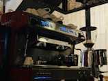 A professional espresso machine with multiple buttons and two portafilters, positioned on a countertop. Several white cups are stacked on top of the machine, which is placed beside a large coffee grinder. The setting suggests a coffee shop environment with a warm and inviting ambiance.