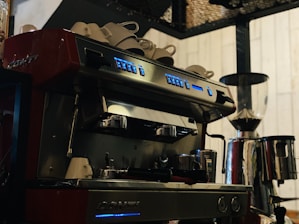 A professional espresso machine with multiple buttons and two portafilters, positioned on a countertop. Several white cups are stacked on top of the machine, which is placed beside a large coffee grinder. The setting suggests a coffee shop environment with a warm and inviting ambiance.