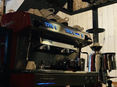 A professional espresso machine with multiple buttons and two portafilters, positioned on a countertop. Several white cups are stacked on top of the machine, which is placed beside a large coffee grinder. The setting suggests a coffee shop environment with a warm and inviting ambiance.