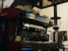 A professional espresso machine with multiple buttons and two portafilters, positioned on a countertop. Several white cups are stacked on top of the machine, which is placed beside a large coffee grinder. The setting suggests a coffee shop environment with a warm and inviting ambiance.