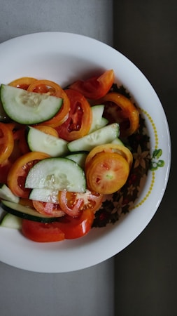A salad comprised of sliced cucumbers and tomatoes is presented in a decorative white bowl with a floral pattern along the edge. The arrangement appears fresh and appetizing, showcasing a mix of vibrant colors.