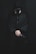 A person dressed in dark clothing, wearing a hood and a face mask, holds a gun with both hands. The setting is dimly lit, adding a mysterious atmosphere. The person has rings on their fingers, and the overall look is tactical or edgy.