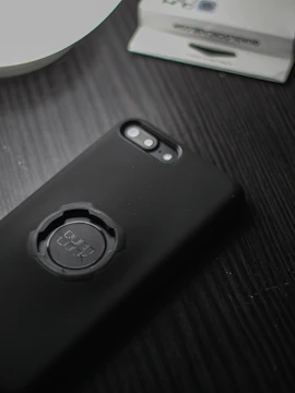 A smartphone with a black case featuring a circular mount labeled 'Quad Lock' is placed on a dark wooden surface. Part of a white dish and a packaging box are visible in the background.