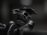 A close-up shot of a mechanical device featuring a hinge and clamp system, primarily made of metallic components. The device appears robust and industrial, positioned against a blurred background that highlights its detailed construction and texture.