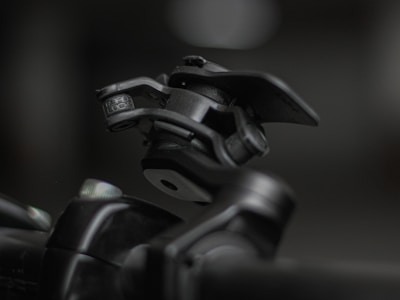 A close-up shot of a mechanical device featuring a hinge and clamp system, primarily made of metallic components. The device appears robust and industrial, positioned against a blurred background that highlights its detailed construction and texture.