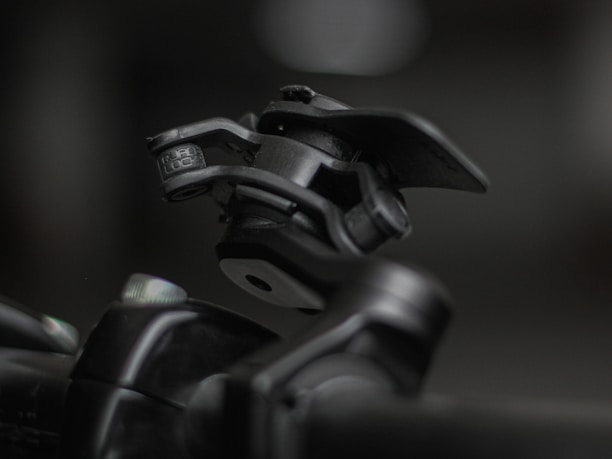 A close-up shot of a mechanical device featuring a hinge and clamp system, primarily made of metallic components. The device appears robust and industrial, positioned against a blurred background that highlights its detailed construction and texture.