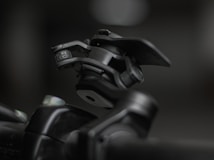 A close-up shot of a mechanical device featuring a hinge and clamp system, primarily made of metallic components. The device appears robust and industrial, positioned against a blurred background that highlights its detailed construction and texture.