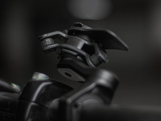 A close-up shot of a mechanical device featuring a hinge and clamp system, primarily made of metallic components. The device appears robust and industrial, positioned against a blurred background that highlights its detailed construction and texture.
