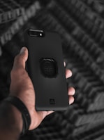 A hand holding a smartphone with a sleek, black protective case. The background features a blurred, textured pattern, creating a contrast with the clear focus on the phone. The phone's camera lens and a grip accessory on the case are clearly visible.