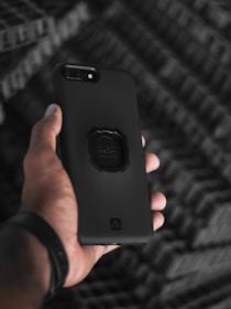 A hand holding a smartphone with a sleek, black protective case. The background features a blurred, textured pattern, creating a contrast with the clear focus on the phone. The phone's camera lens and a grip accessory on the case are clearly visible.