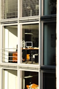 A modern office building with large glass windows reflecting sunlight. Inside, there are desks with computers and office equipment. The scene is quiet and orderly, suggesting it might be outside of regular working hours.