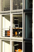 A modern office building with large glass windows reflecting sunlight. Inside, there are desks with computers and office equipment. The scene is quiet and orderly, suggesting it might be outside of regular working hours.