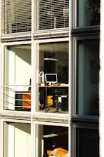 A modern office building with large glass windows reflecting sunlight. Inside, there are desks with computers and office equipment. The scene is quiet and orderly, suggesting it might be outside of regular working hours.