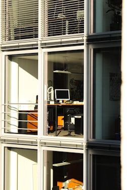 A modern office building with large glass windows reflecting sunlight. Inside, there are desks with computers and office equipment. The scene is quiet and orderly, suggesting it might be outside of regular working hours.