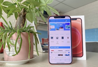 A smartphone with a red case is displayed in front of its packaging box. A website is open on the screen, showing various tech products for sale. Next to the phone, there is a potted plant with green leaves in a decorative pink pot placed on a saucer. The background includes a blurred interior setting with walls and what appears to be another electronic device.