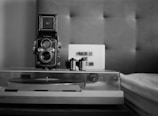 A vintage twin-lens reflex camera is prominently placed on a surface, accompanied by two rolls of film. In the background, a sign with the text 'PROCESS OR ABANDON' is visible. The entire scene is set in a monochrome, retro style with a turntable partially visible at the bottom.