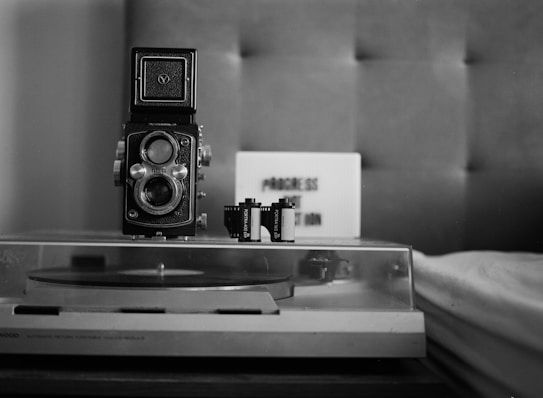 A vintage twin-lens reflex camera is prominently placed on a surface, accompanied by two rolls of film. In the background, a sign with the text 'PROCESS OR ABANDON' is visible. The entire scene is set in a monochrome, retro style with a turntable partially visible at the bottom.