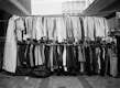 A street market display with two racks of assorted clothing. The top rack has shirts of various styles and patterns, while the bottom rack features pants and blazers. The setting appears to be an outdoor urban environment with buildings in the background.