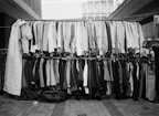 A street market display with two racks of assorted clothing. The top rack has shirts of various styles and patterns, while the bottom rack features pants and blazers. The setting appears to be an outdoor urban environment with buildings in the background.