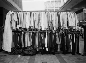 A street market display with two racks of assorted clothing. The top rack has shirts of various styles and patterns, while the bottom rack features pants and blazers. The setting appears to be an outdoor urban environment with buildings in the background.