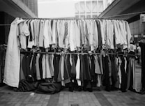 A street market display with two racks of assorted clothing. The top rack has shirts of various styles and patterns, while the bottom rack features pants and blazers. The setting appears to be an outdoor urban environment with buildings in the background.