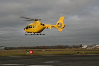 A yellow air ambulance helicopter is hovering over an airfield against a backdrop of overcast skies. The helicopter's rotors are spinning, and it has the markings of an emergency service vehicle. The landscape features grass, a tarmac area, and some buildings in the distance.