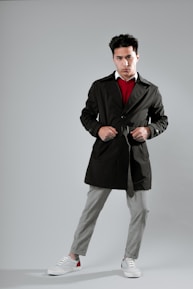 A man is standing confidently with his hands on the belt of a dark trench coat. He is wearing a red sweater, a white shirt underneath, gray plaid trousers, and casual sneakers. The background is a neutral gray, highlighting his fashion-forward look.